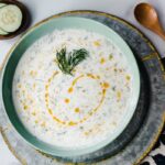 bowl of yogurt soup with dill and cucumbers on the sides